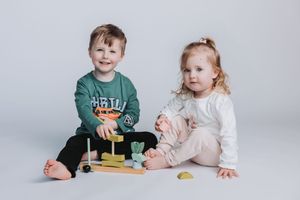 Two young toddlers sitting on white background playing with colorful wooden stacking toy together.