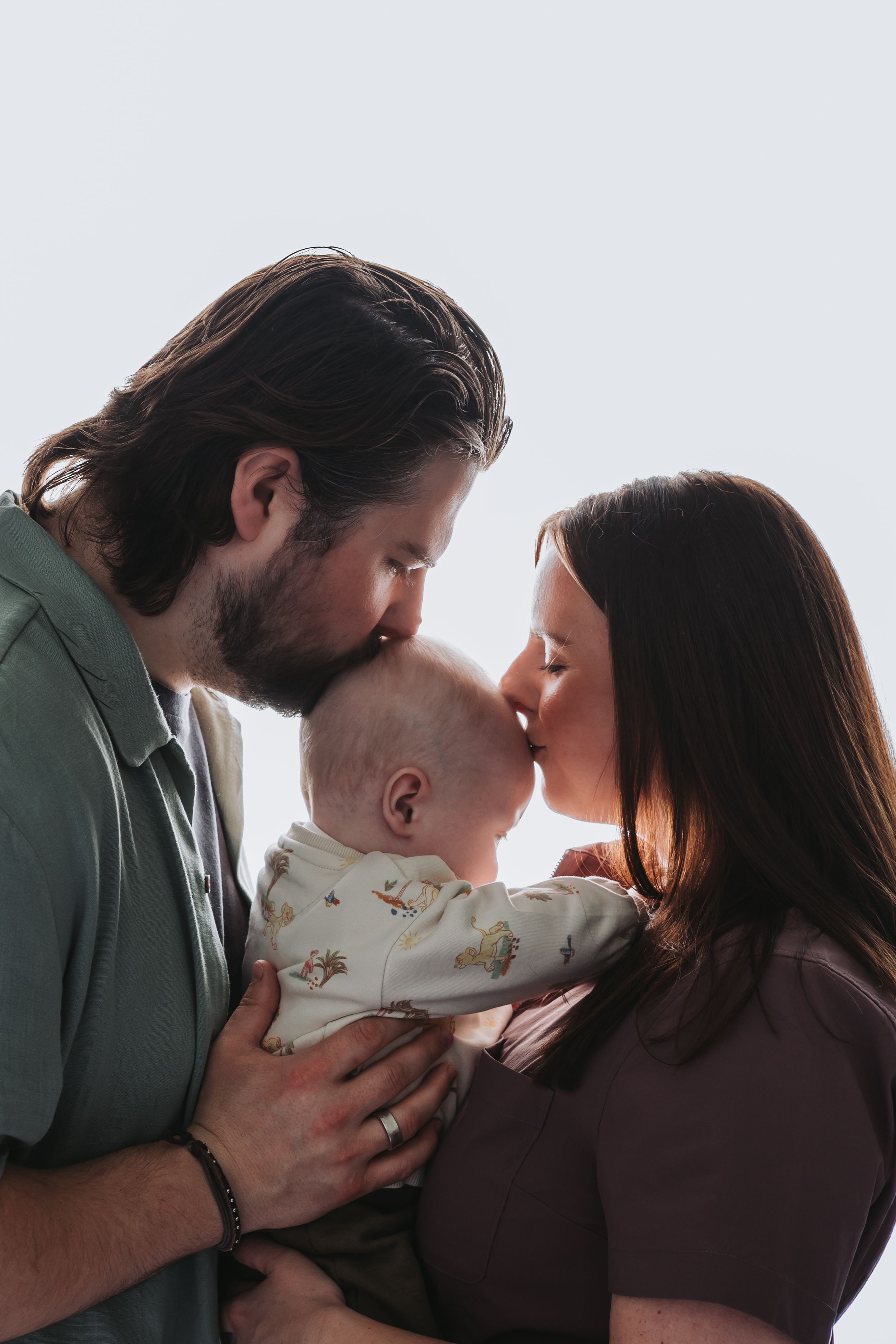 A couple shares a kiss while holding their newborn baby between them in a tender family portrait.