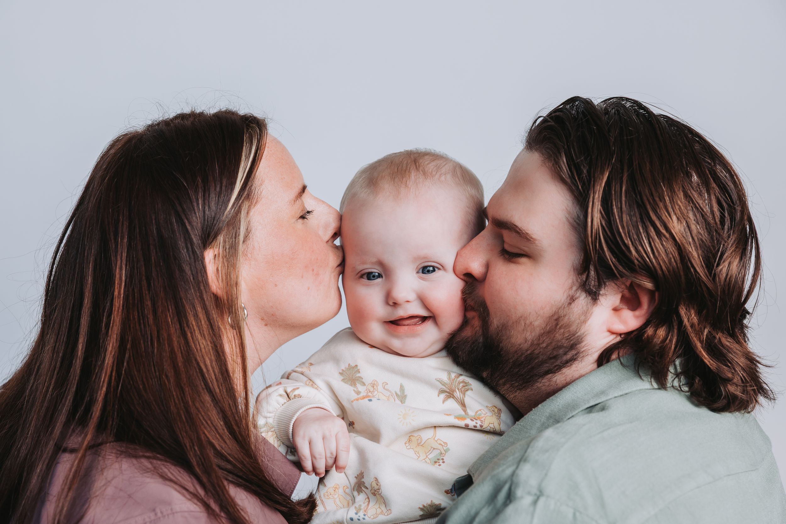 Happy baby smiling while parents give kisses on cheeks in a sweet family portrait photo.