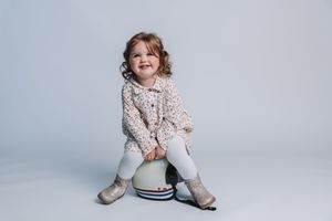 Smiling toddler girl with curly hair sitting on a helmet, wearing a floral jacket and white leggings.