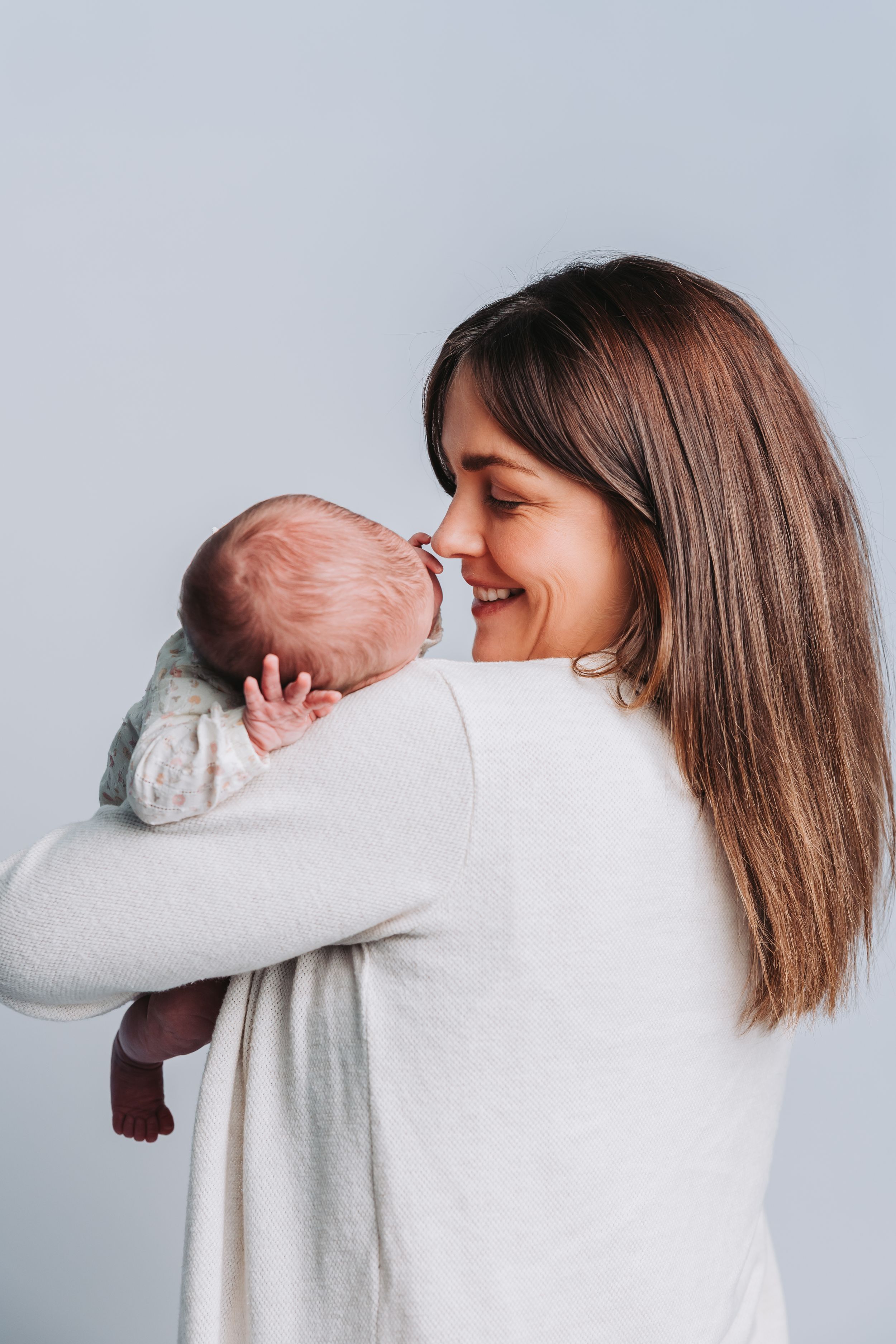 Smiling mother holding newborn baby close to her face, wearing white sweater against light blue background.