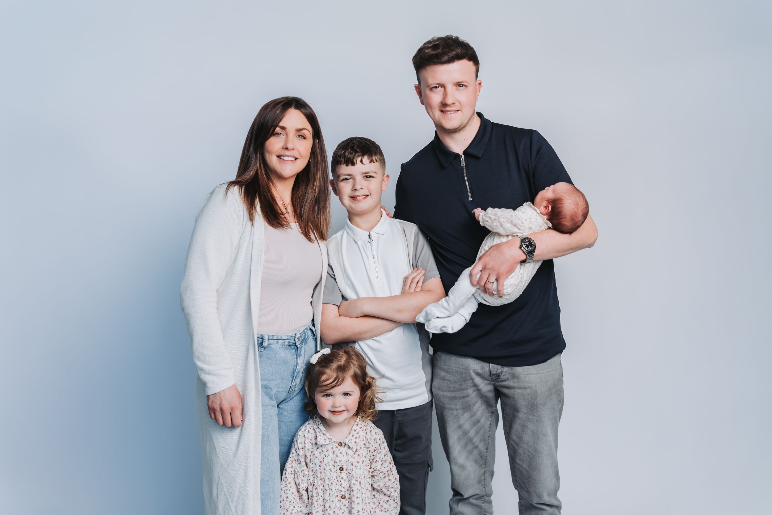 Family of five posing together, parents with two children and a newborn baby, against a light blue studio background.