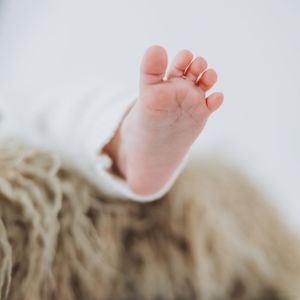 Close-up of a newborn baby's tiny foot resting on a fluffy beige blanket with small gold rings on toes.
