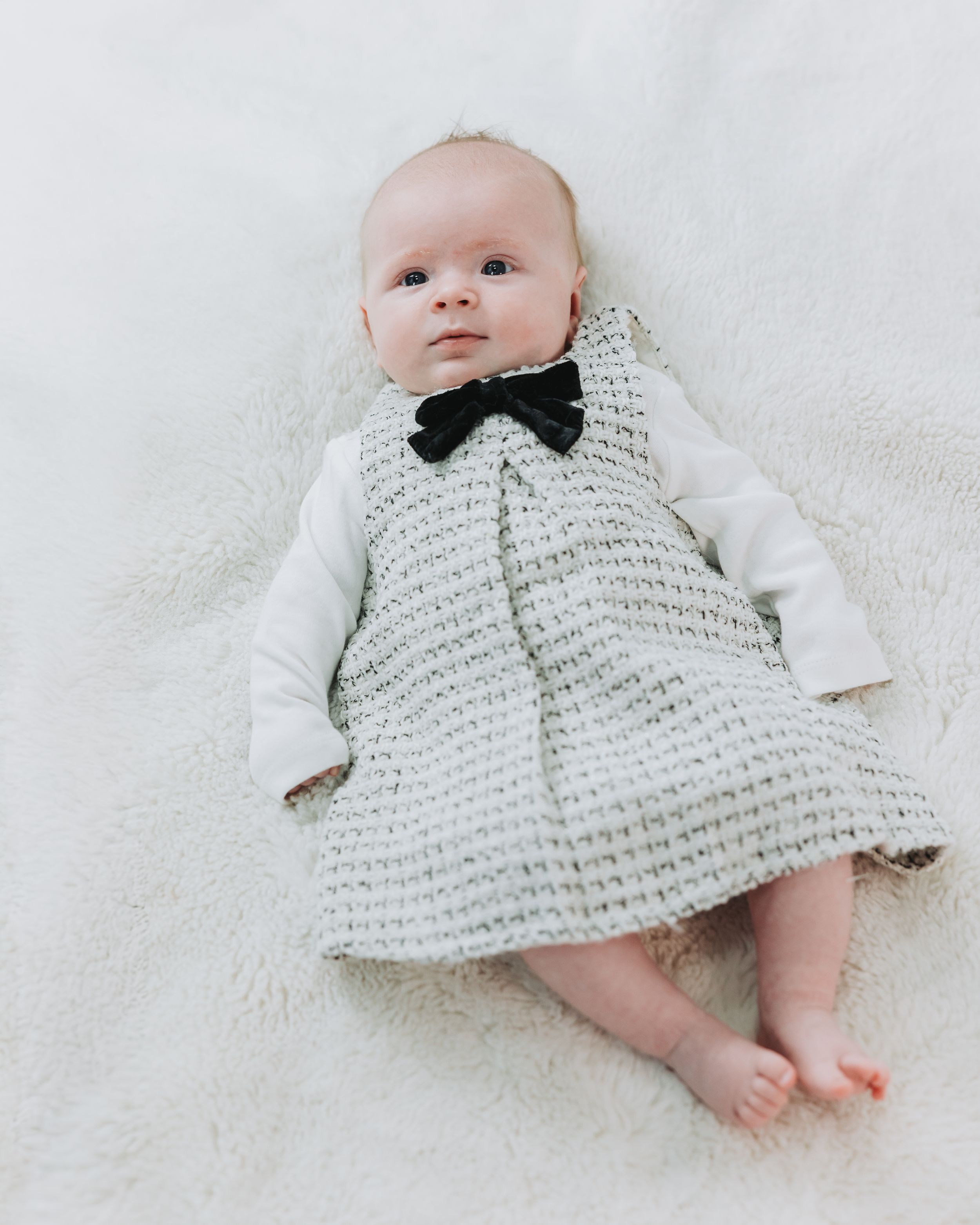 Baby in knit dress with black bow tie lying on fluffy white blanket, looking up curiously.