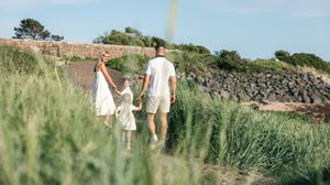 Family of three walking hand-in-hand along a scenic coastal path surrounded by lush green grass and stone walls.