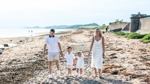 Family of four in white outfits walking hand-in-hand on a scenic sandy beach with historic stone walls.