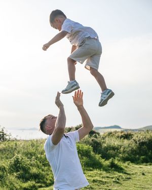 A father joyfully tosses his young son high into the air outdoors on a sunny day near a green hillside.