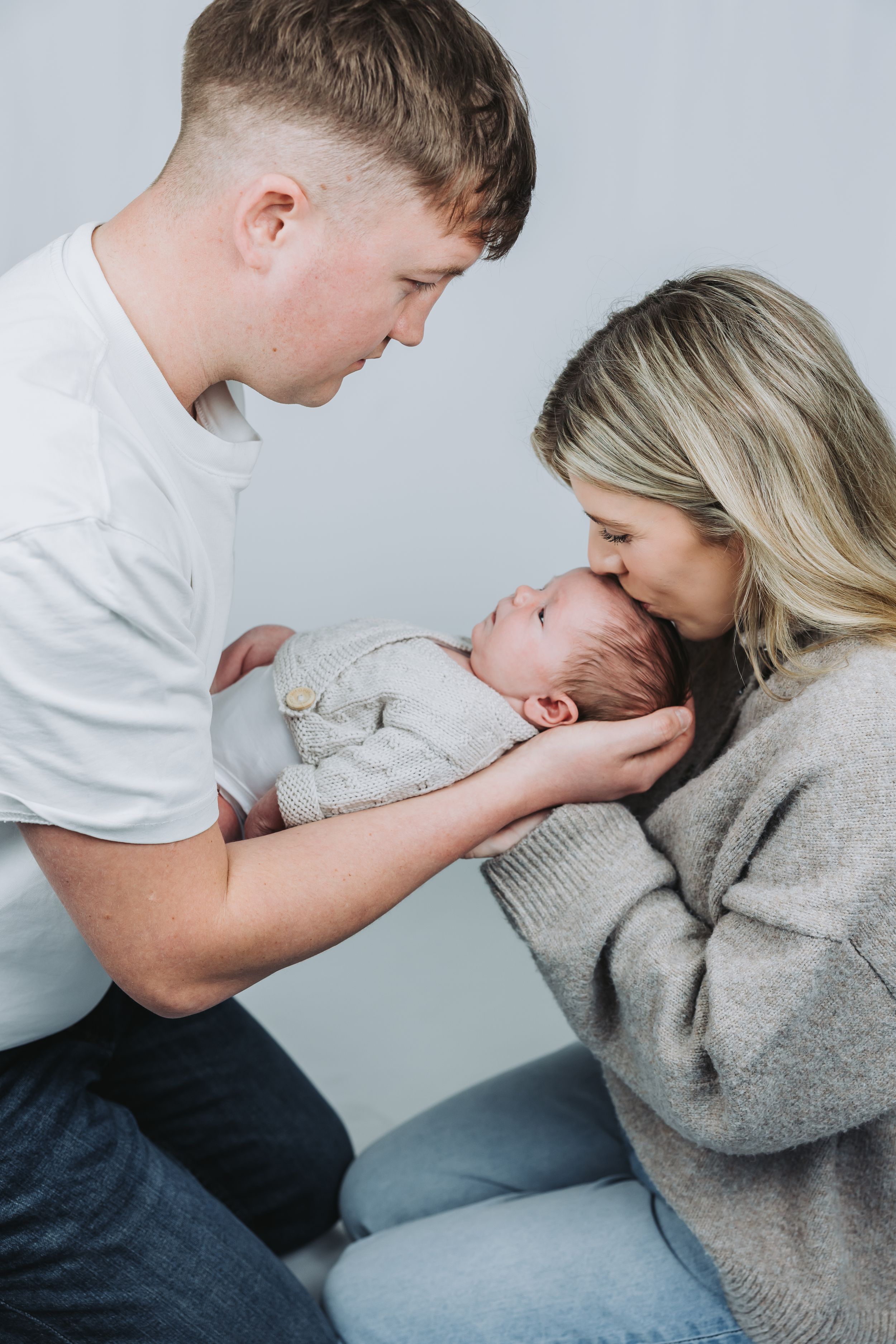 Young parents lovingly gazing at their newborn baby, mother kissing infant while father gently touches the child.