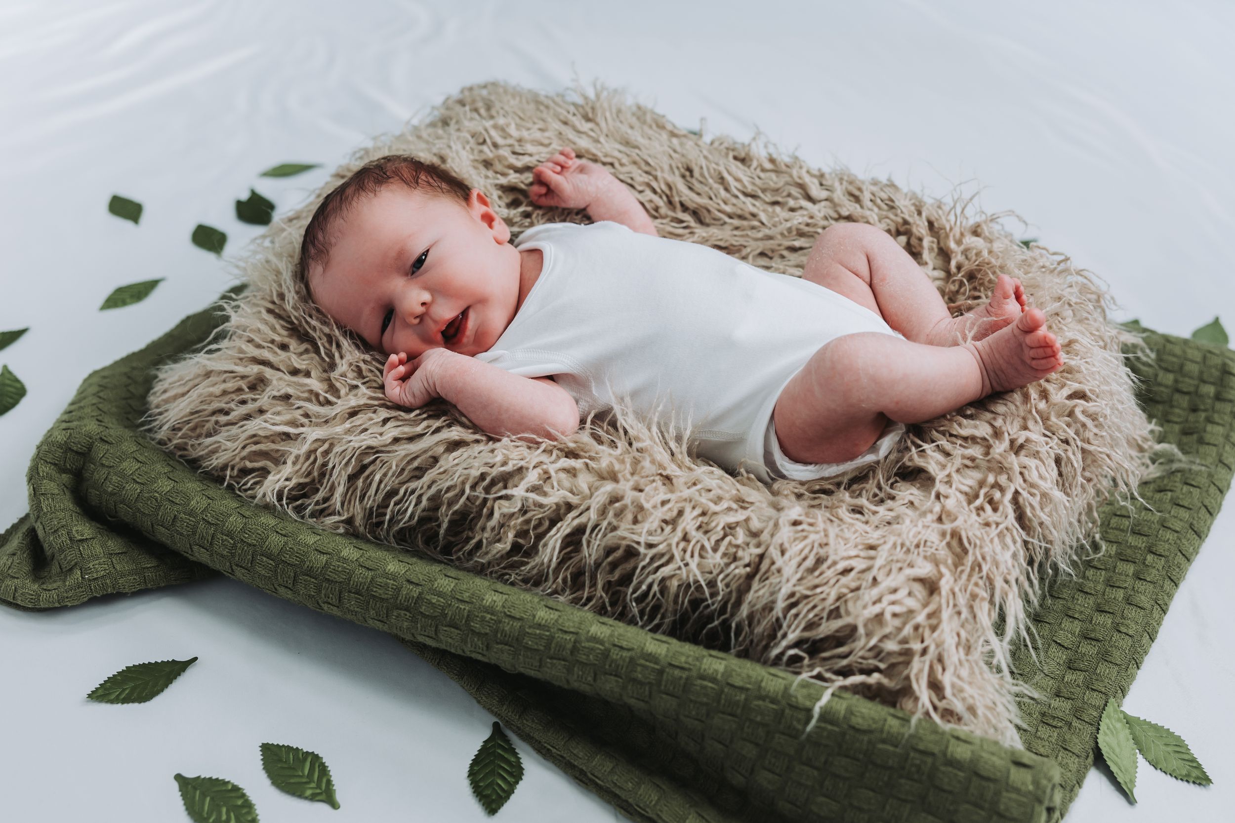 Newborn baby in white onesie sleeping peacefully on fluffy beige fur blanket atop green knit mat with leaf decorations.