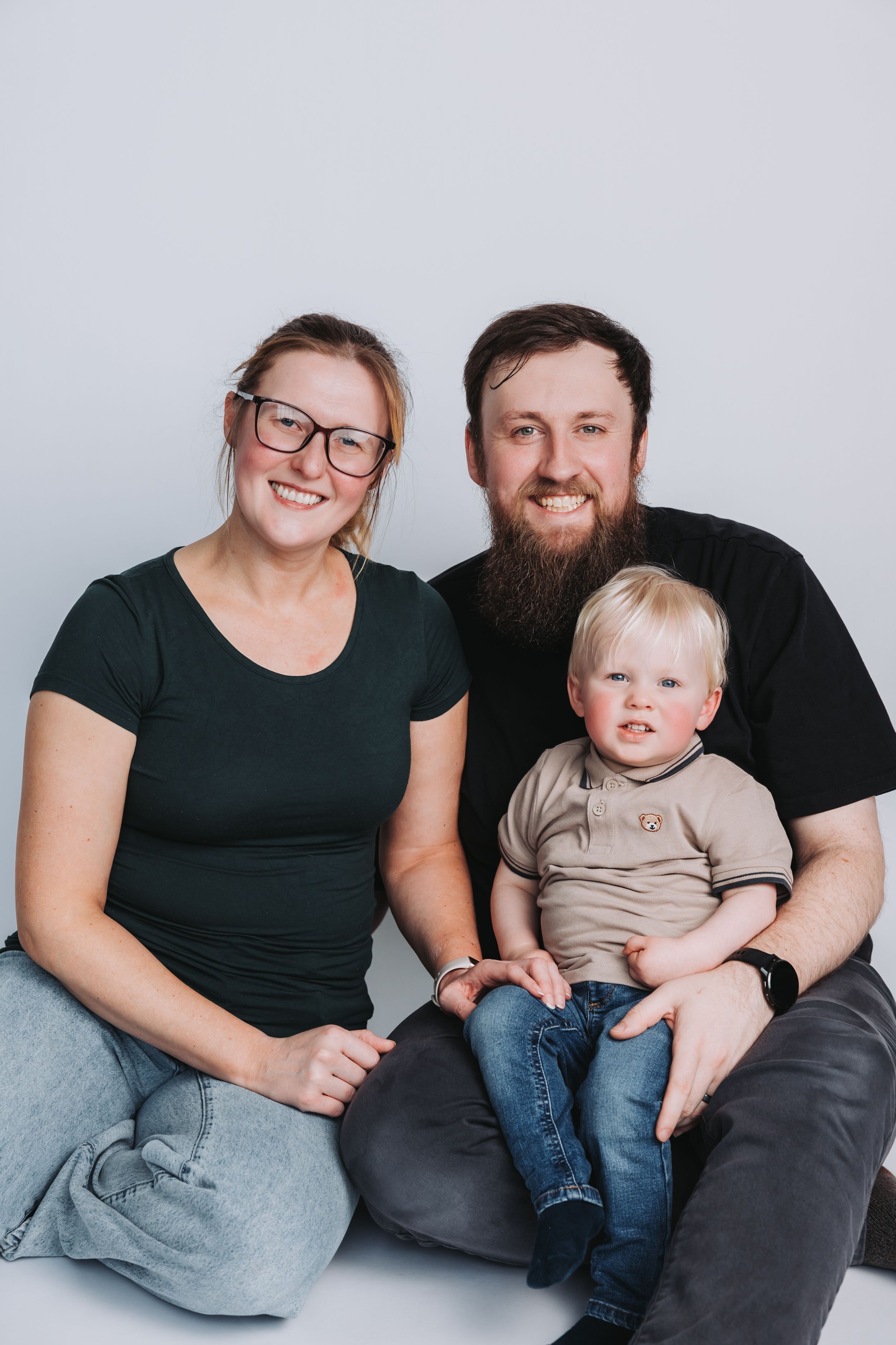 A smiling family of three poses in a studio, with a bearded man, woman in glasses, and a blonde toddler boy.