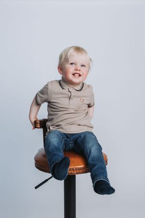 Smiling blonde toddler boy sitting on a brown bar stool, wearing a beige polo shirt and jeans.