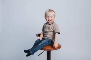 Smiling blonde toddler boy sitting on a brown bar stool wearing jeans and a gray t-shirt against a white background.
