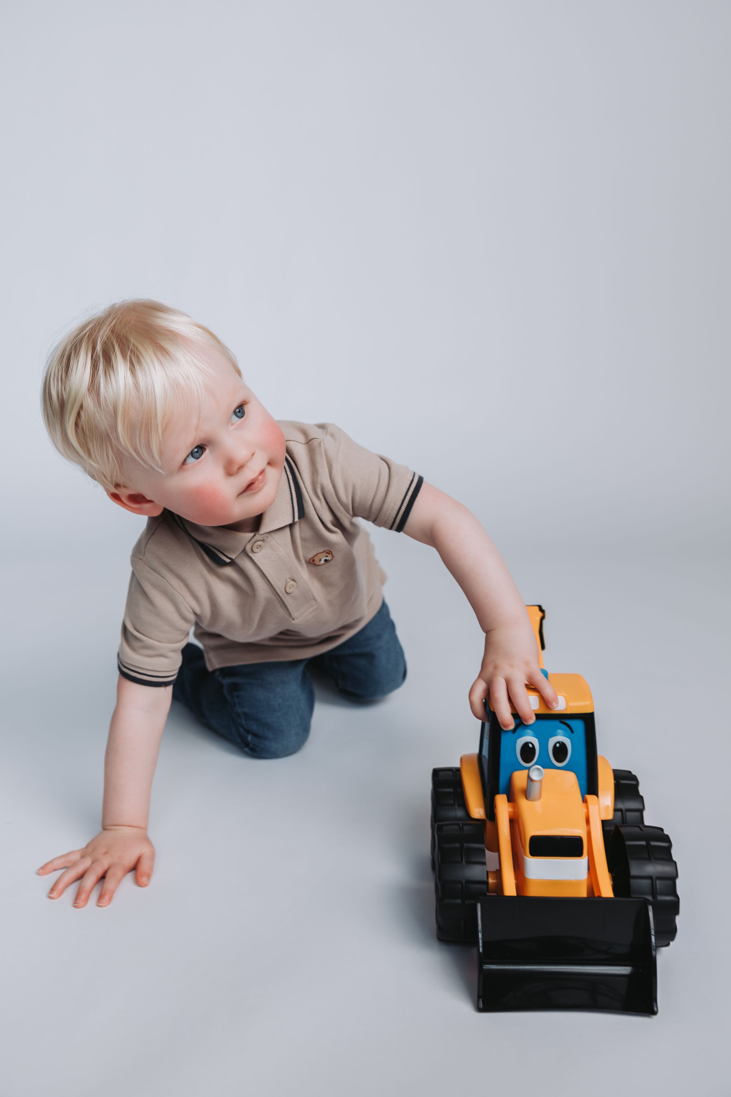 Toddler boy with blonde hair playing with an orange toy bulldozer truck on a white studio background.