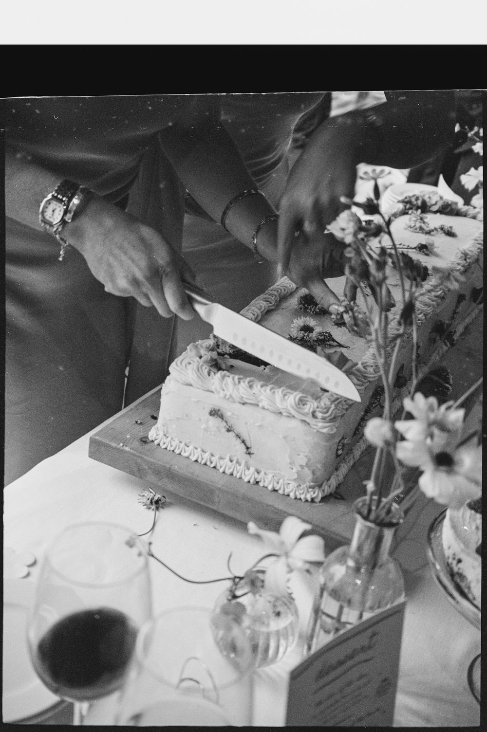 Black and white photo of hands cutting a decorated celebration cake on a table with flowers and glassware.