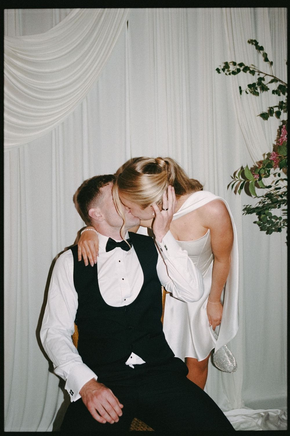 A bride and groom share a romantic kiss at their wedding reception, with white draping and floral arrangements in the background.