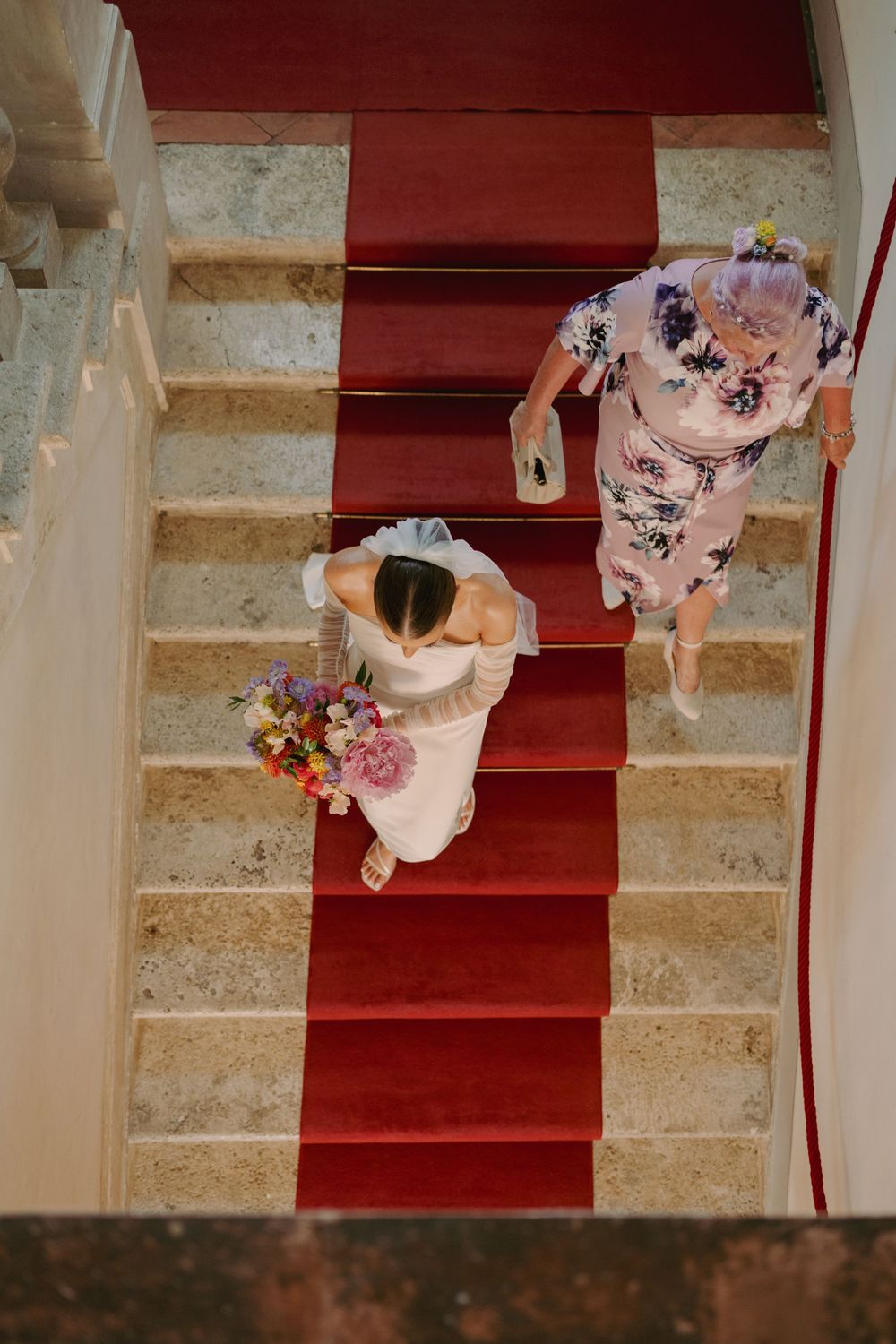 Aerial view of a bride in white dress holding colorful bouquet ascending red-carpeted stairs with a guest in floral dress.