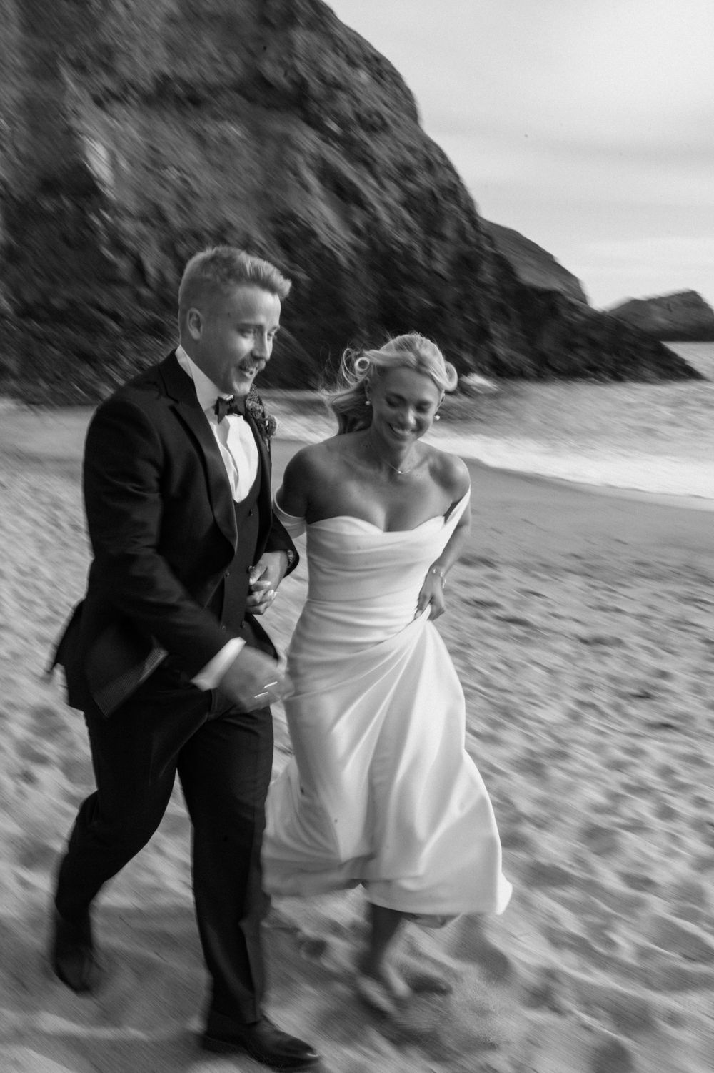 Black and white photo of a bride and groom laughing while running on a sandy beach, rocks in background.