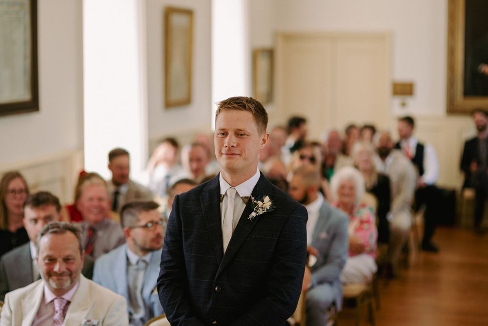 Groom in navy suit with boutonniere stands at altar awaiting bride, wedding guests seated in elegant ceremony hall.