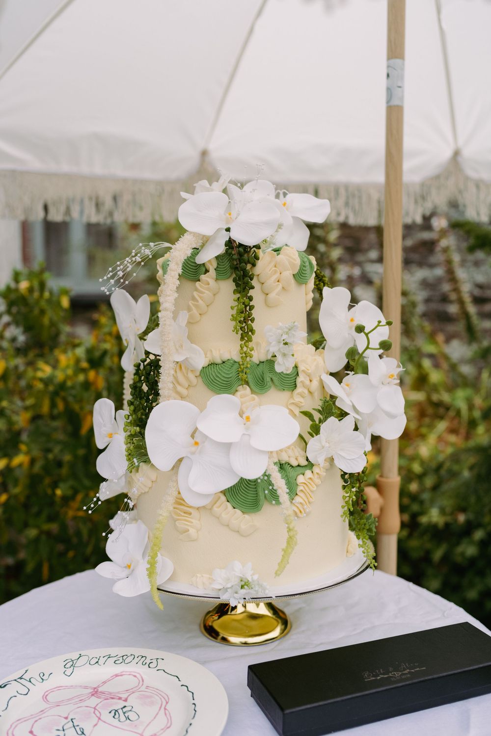 Elegant three-tier white wedding cake adorned with white orchids and green foliage on a gold stand outdoors.