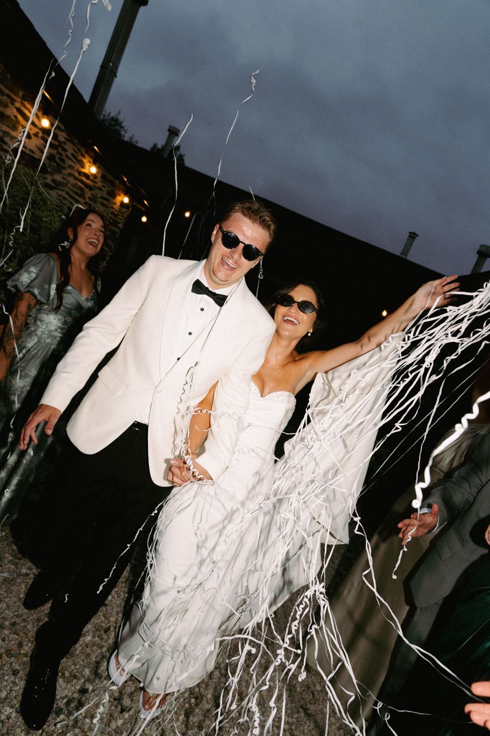 A bride and groom in white tuxedo and wedding gown celebrate with silver streamers at an outdoor evening wedding reception.
