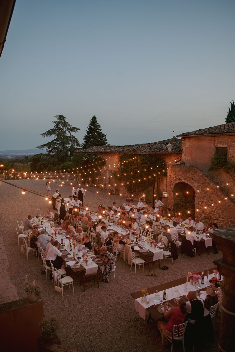 Outdoor wedding reception at dusk with string lights over long dining tables at a rustic Tuscan villa.
