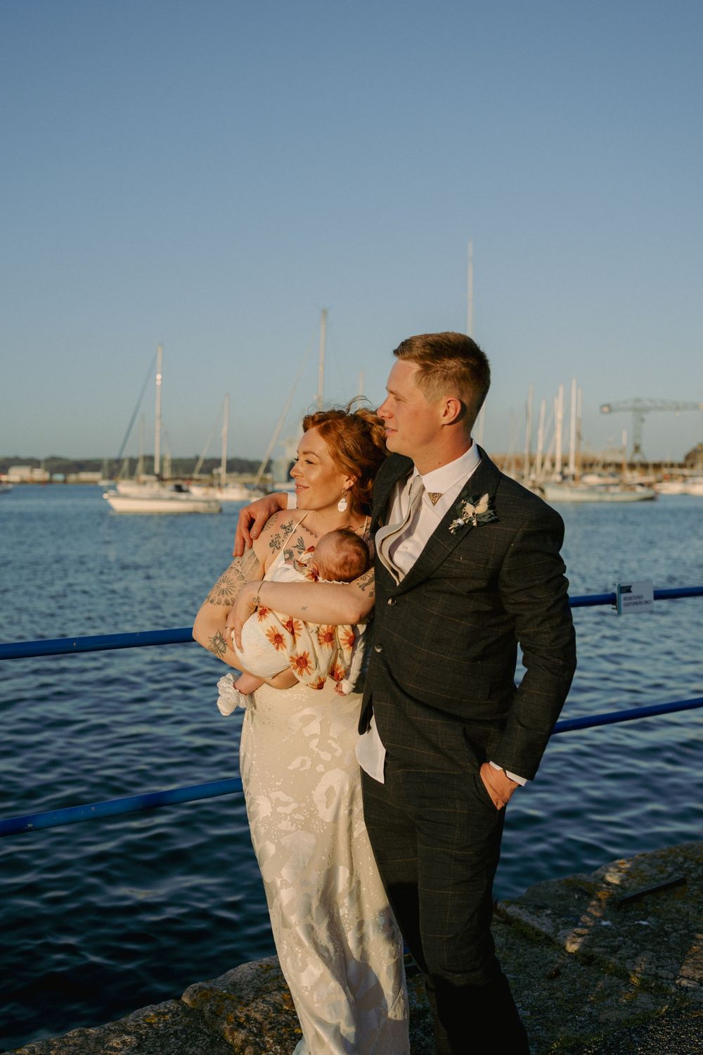 Bride and groom holding newborn baby by waterfront marina on their wedding day in golden hour light.