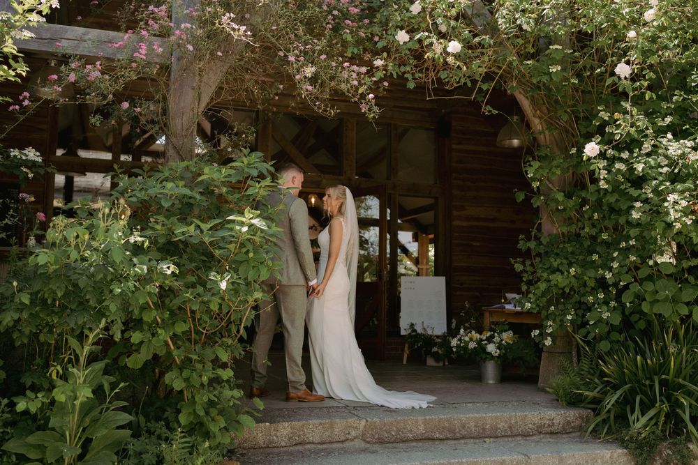 Couple holding hands at rustic barn wedding venue surrounded by lush greenery and blooming flowers.