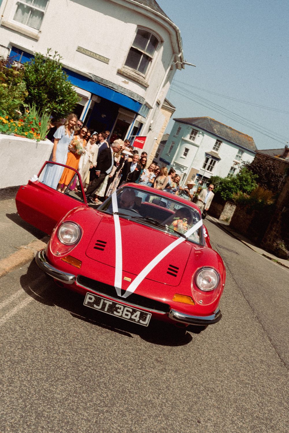 Red Ferrari Dino wedding car with white ribbon drives through a quaint village street with guests gathered outside.