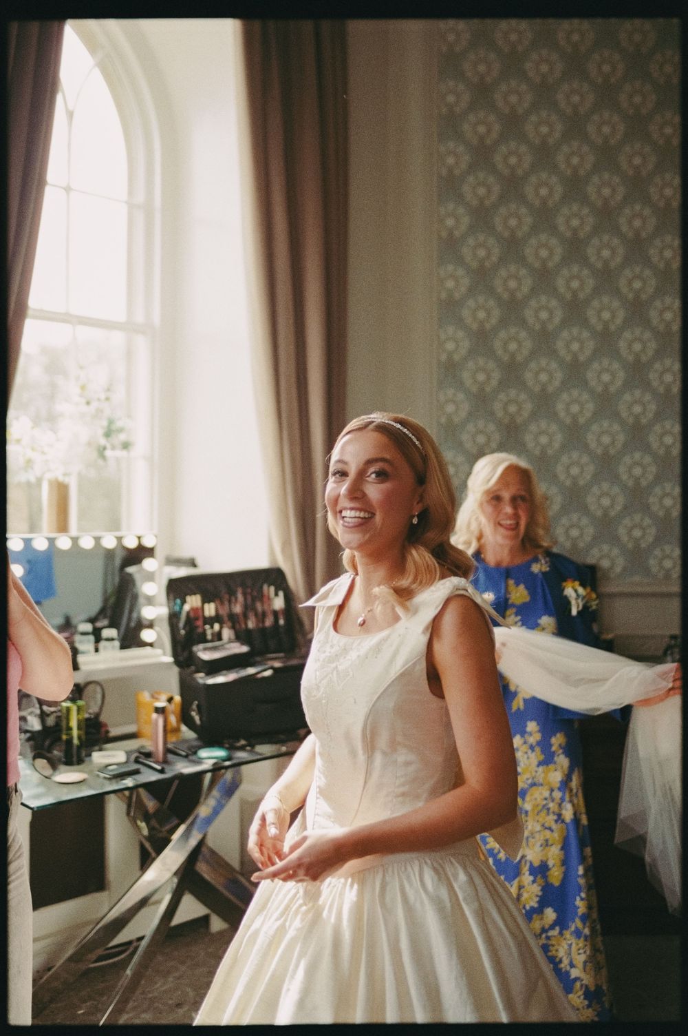 A smiling bride in a white gown stands in an elegant dressing room while a woman in blue adjusts her dress.