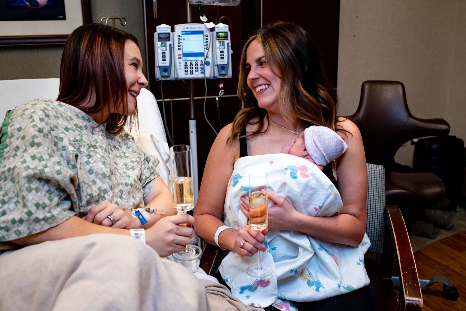 Two new parents share champagne toasts in a hospital room while one holds a newborn baby wrapped in a blanket.