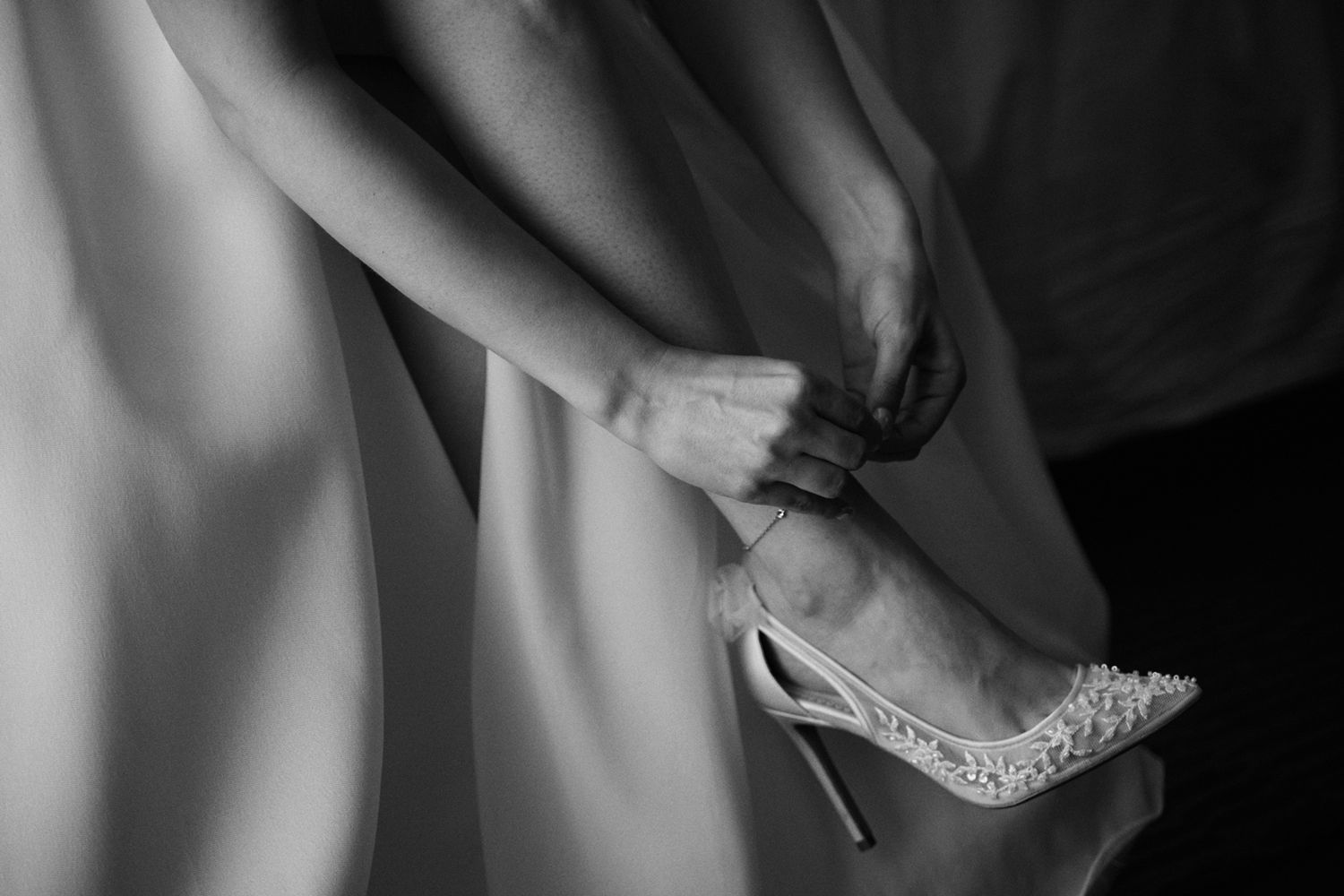 Black and white photo of a bride fastening a pearl-embellished stiletto heel on her foot beneath a flowing gown.