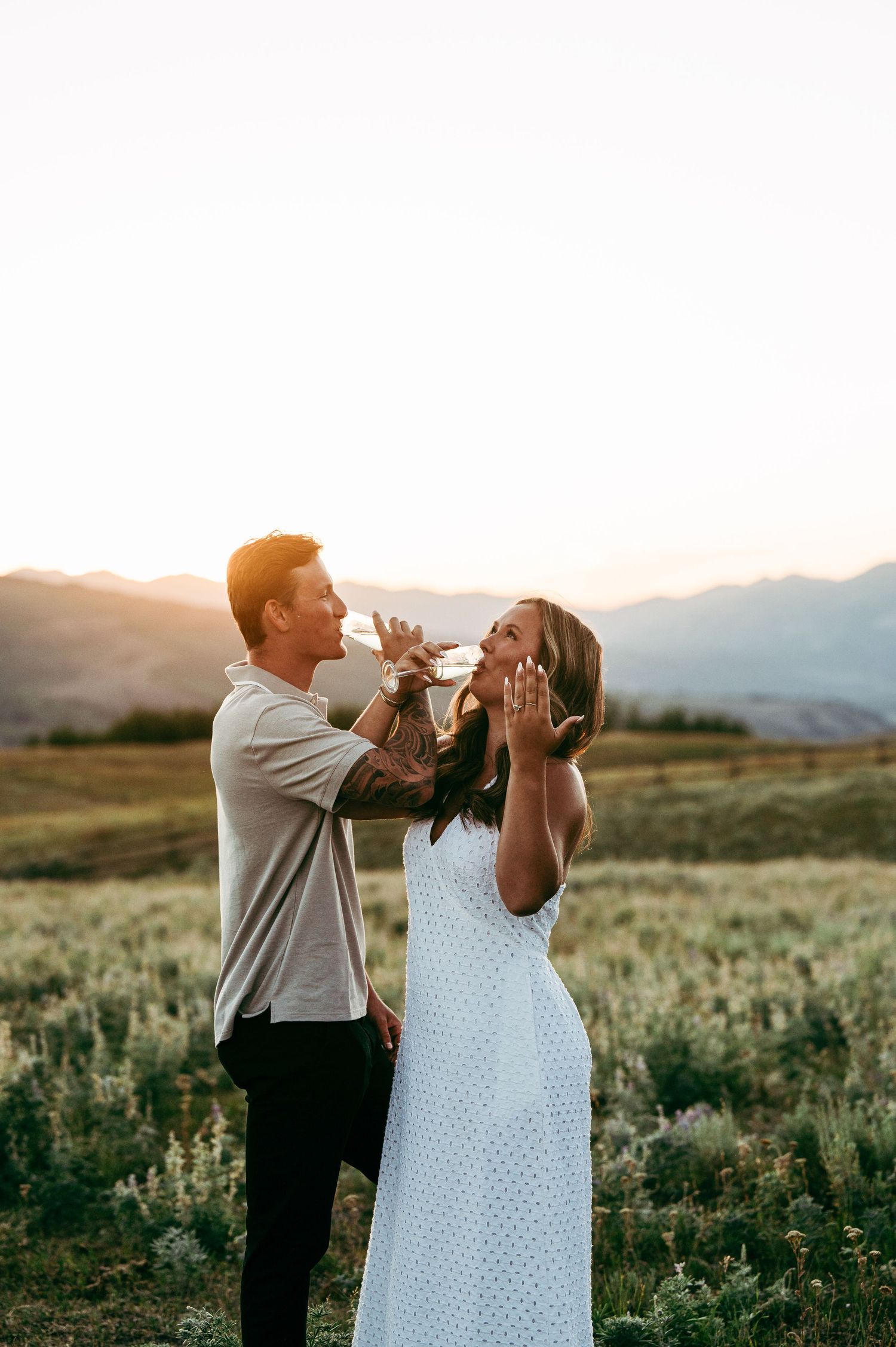 Couple sharing a romantic moment at golden hour in a mountain meadow, woman showing engagement ring.