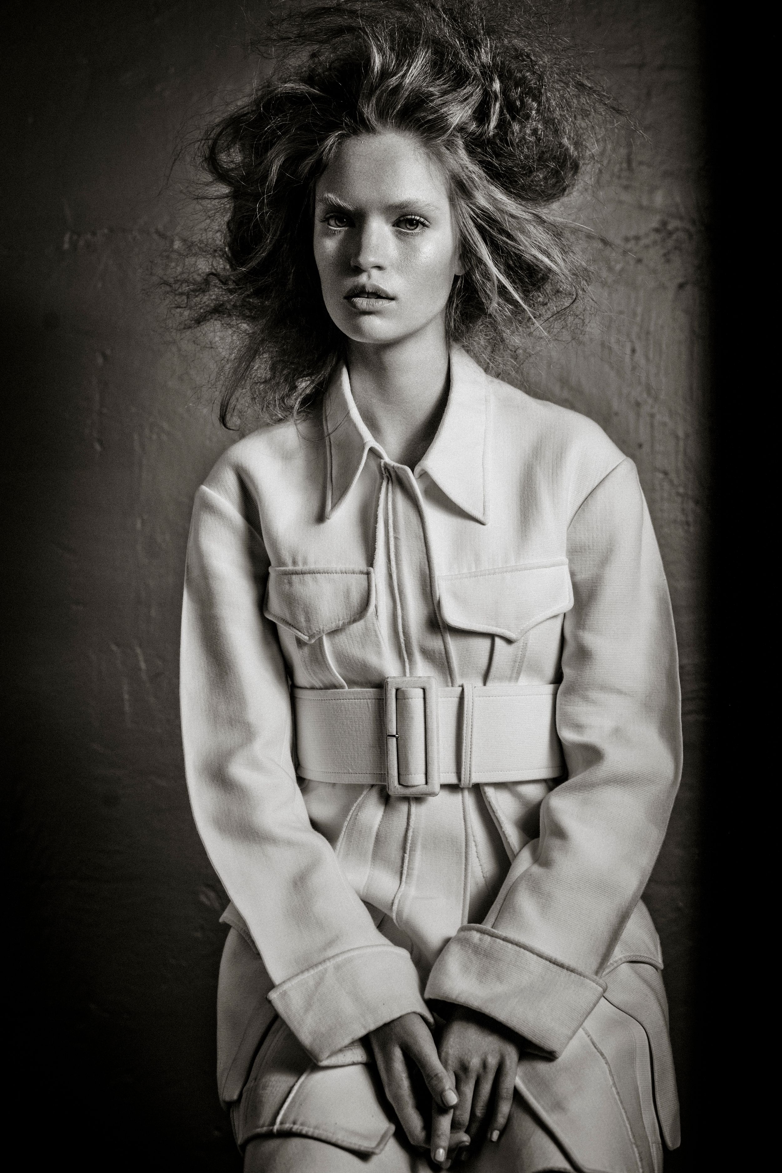 Black and white fashion photo of a woman in a belted utility jacket with windswept hair against a dark background.