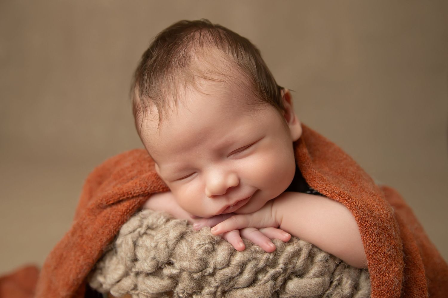 Smiling sleeping newborn baby wrapped in orange blanket posed in bowl for studio photography session.