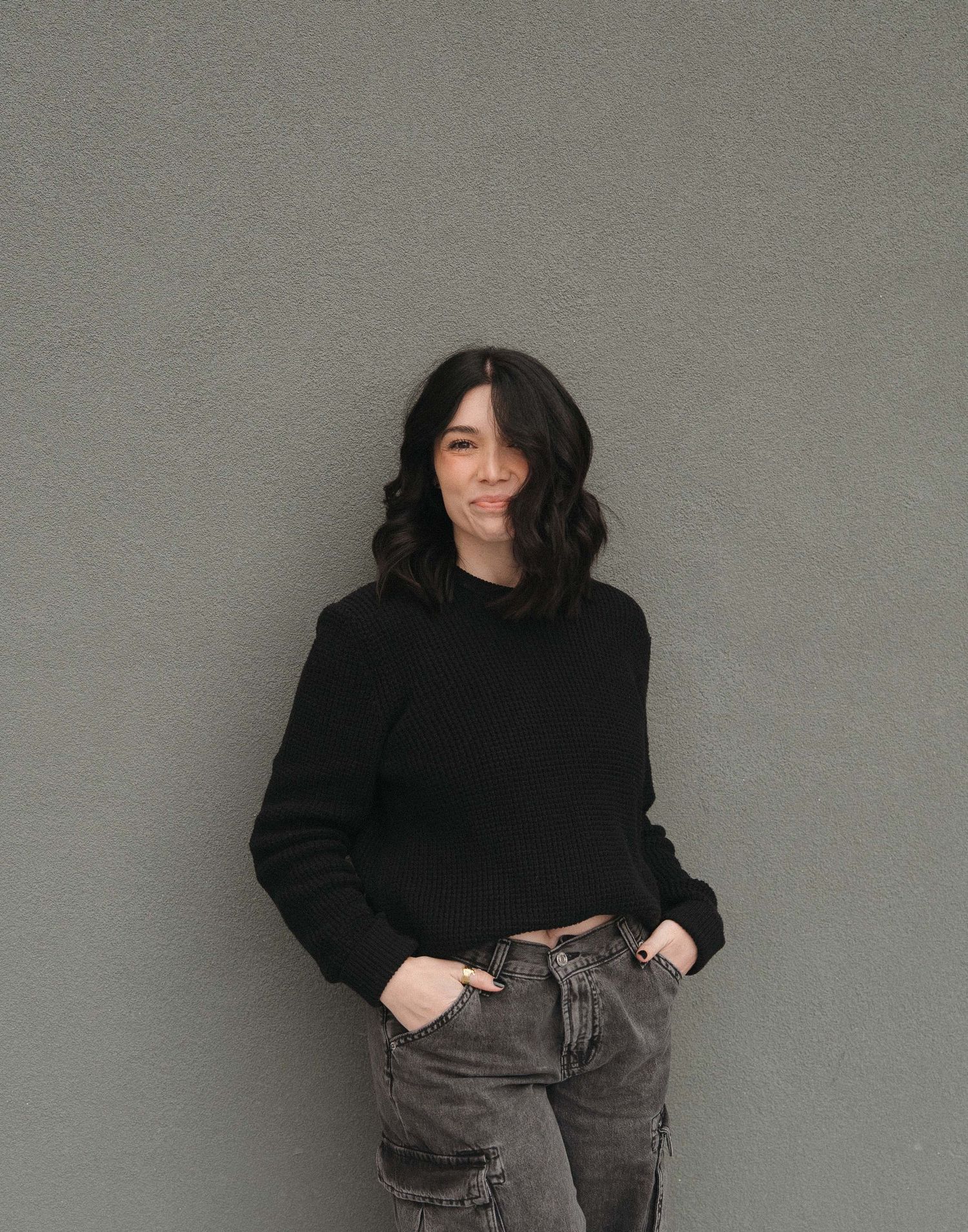 Destination wedding photographer with dark hair wearing a black cropped sweater and gray cargo jeans posing against a gray wall.