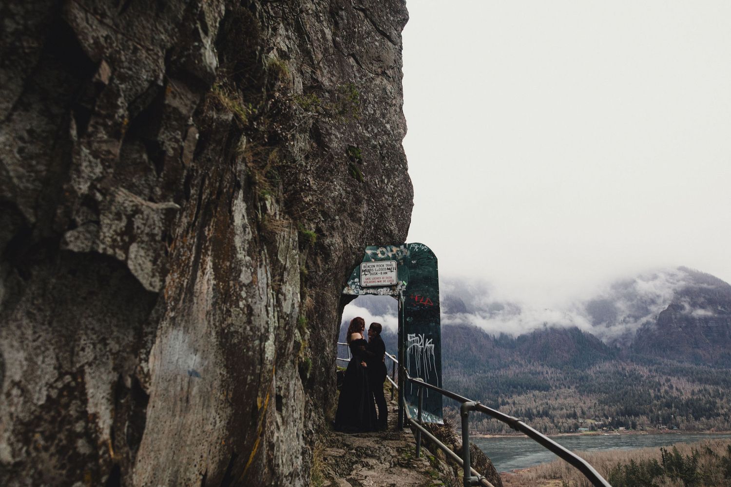 Couple standing beneath a dramatic rocky cliff overhang at a scenic mountain overlook on a cloudy day.