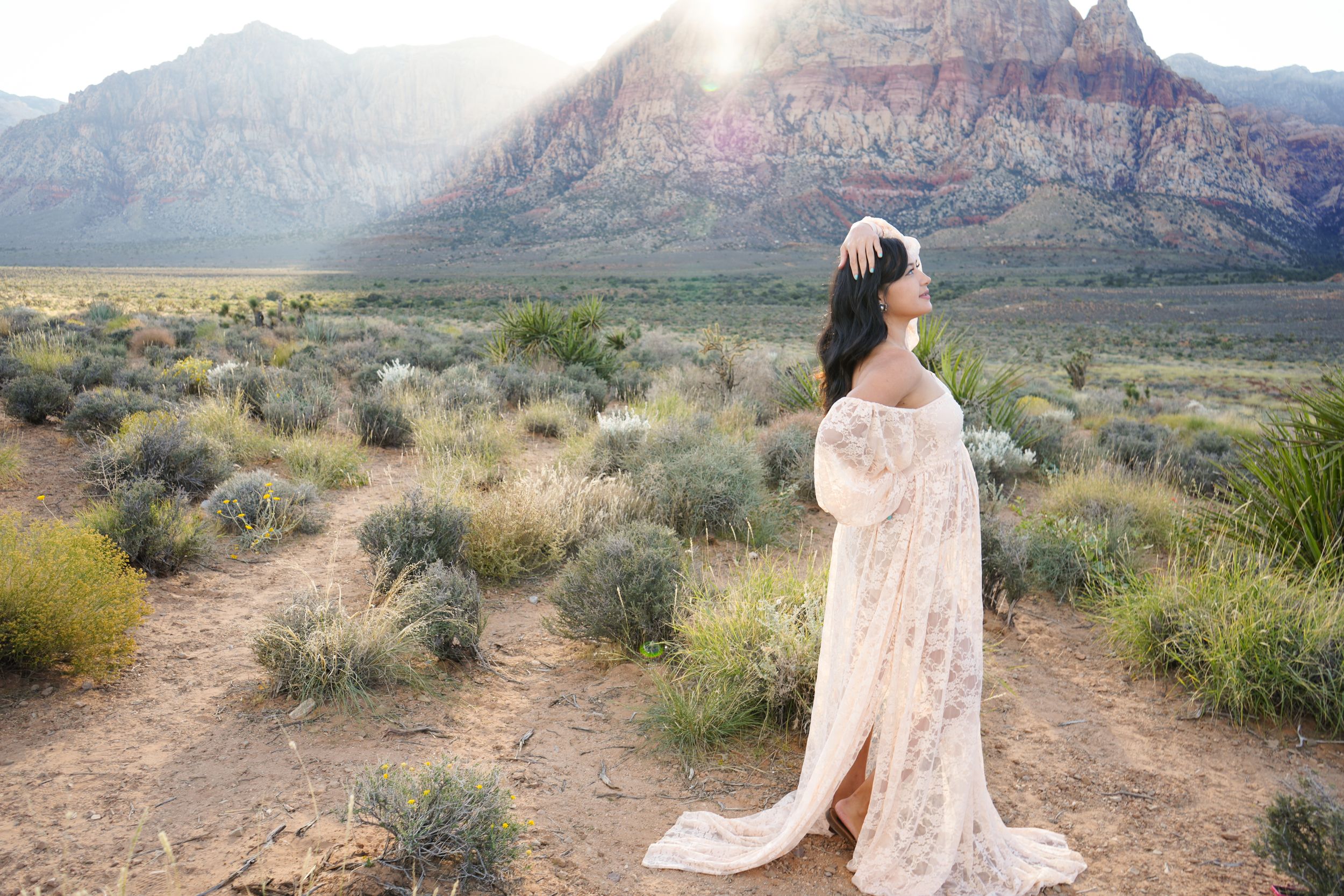 Woman in lace gown poses in desert landscape with red rock mountains and golden sunlight behind her.