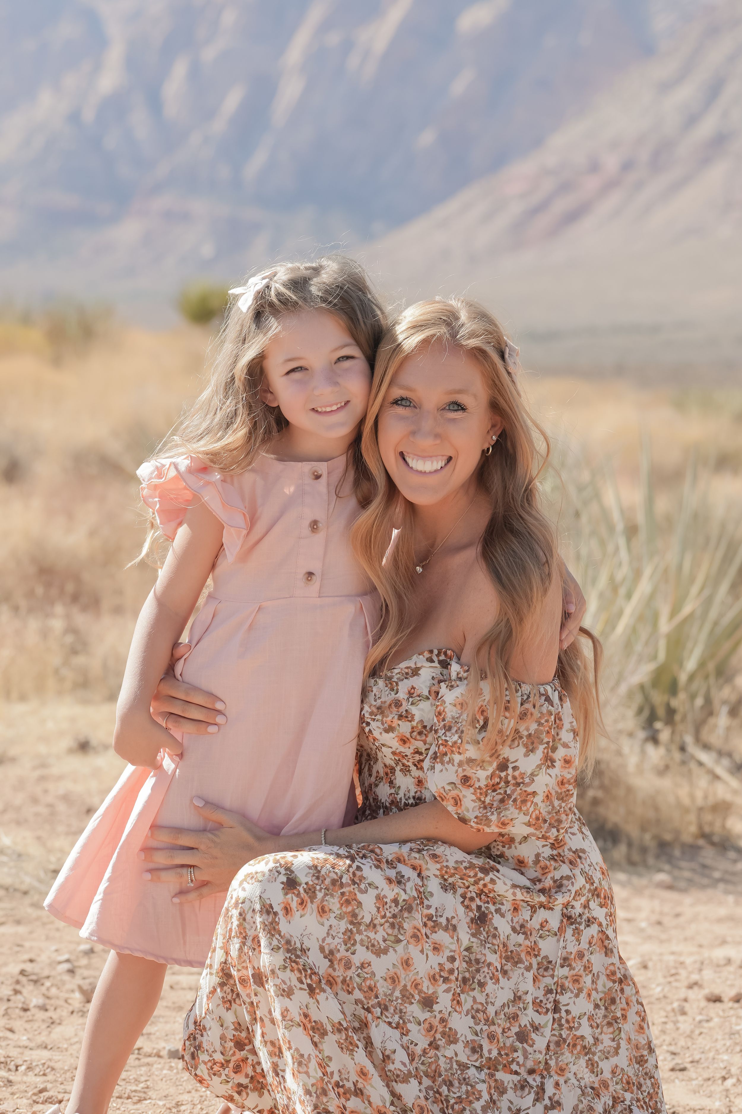 Smiling blonde woman in floral dress posing with young girl in pink dress in desert landscape.