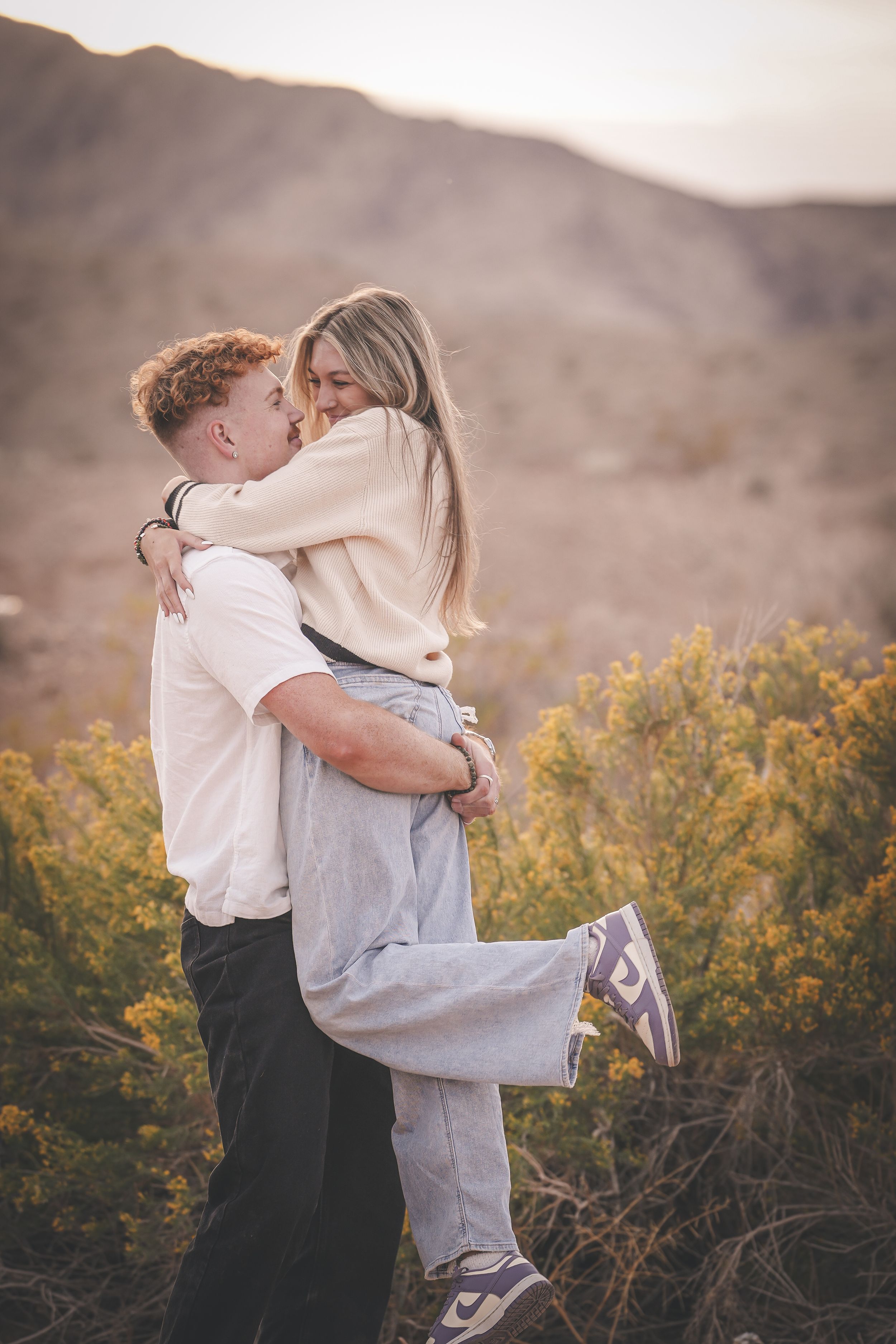 Couple sharing a romantic embrace outdoors with desert mountains and yellow wildflowers in the background.