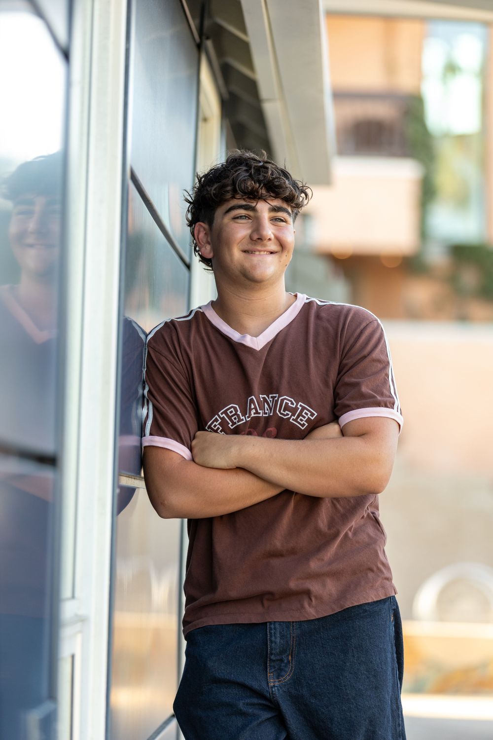 Smiling teenage boy in a brown France t-shirt leaning against a glass window with arms crossed outdoors.
