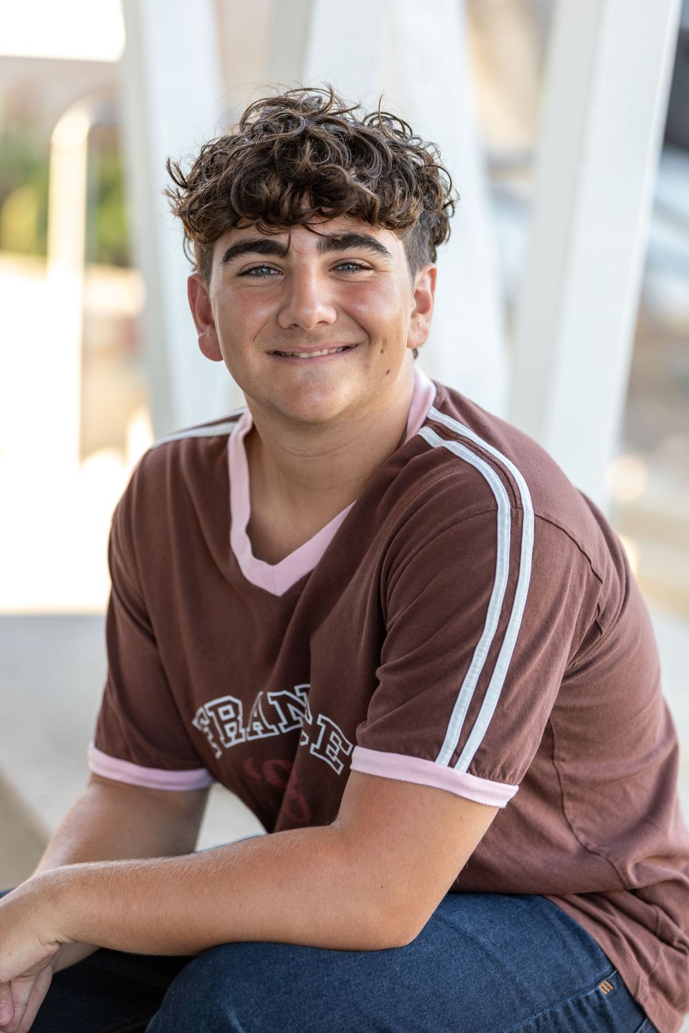 Smiling teenage boy with curly hair wearing a brown jersey shirt, sitting outdoors in a casual portrait photo.