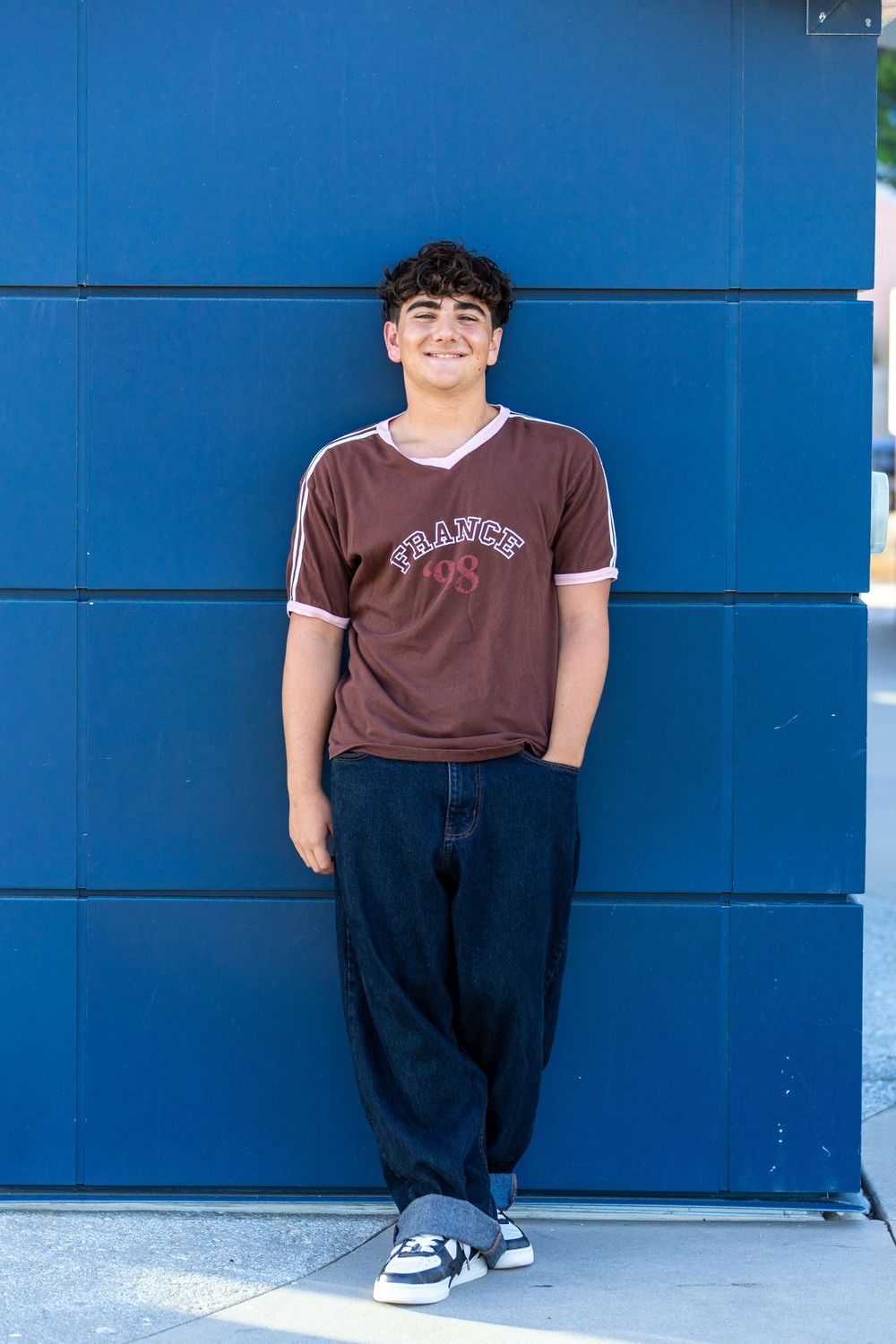 Smiling teen boy in brown France t-shirt and baggy jeans leaning against a blue wall outdoors.