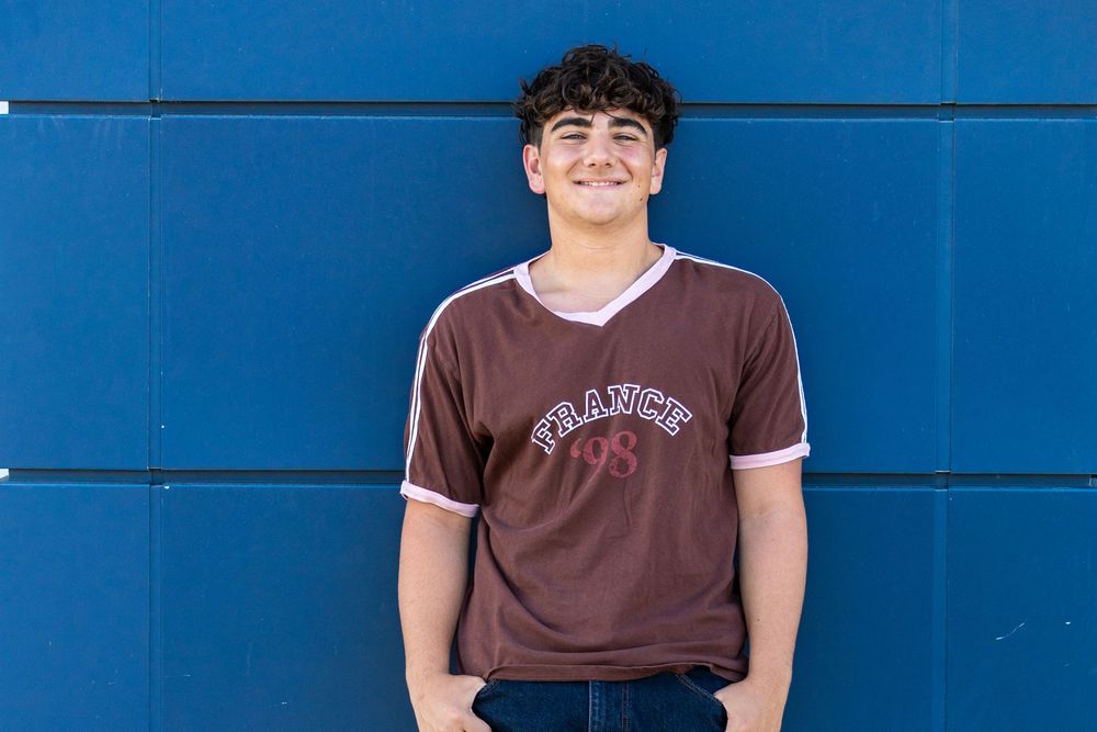 Smiling teenage boy in brown France jersey standing against a blue wall outdoors.