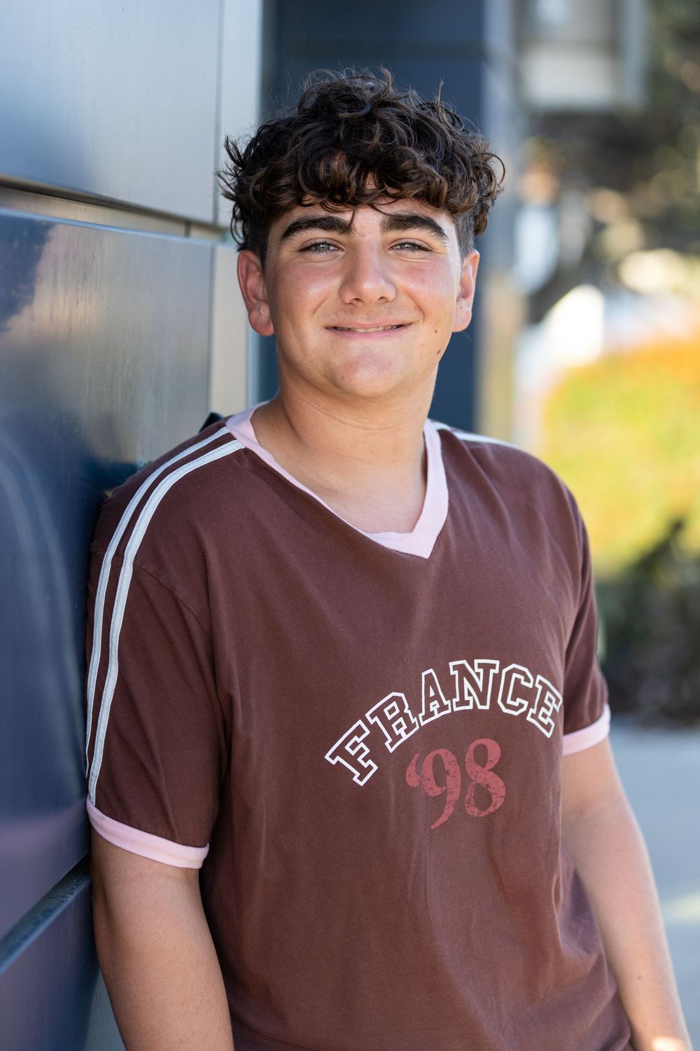 Smiling teenage boy with curly hair wearing a brown France 98 soccer jersey leaning against a wall.