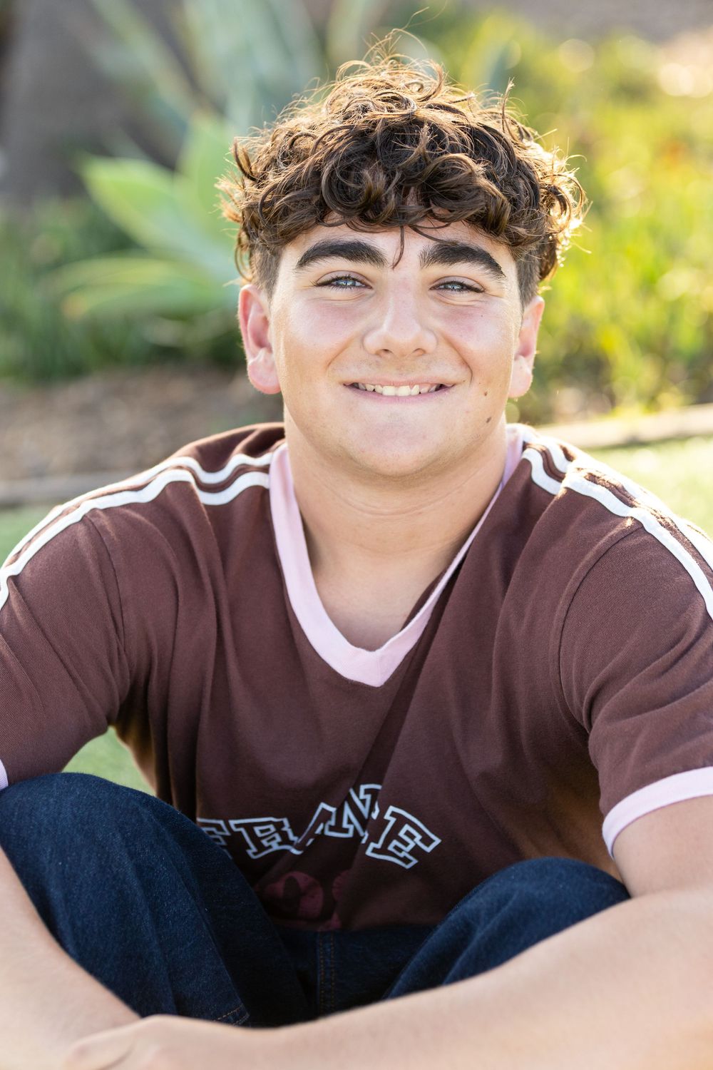 Smiling teenage boy with curly hair wearing a maroon jersey, sitting cross-legged outdoors in natural sunlight.