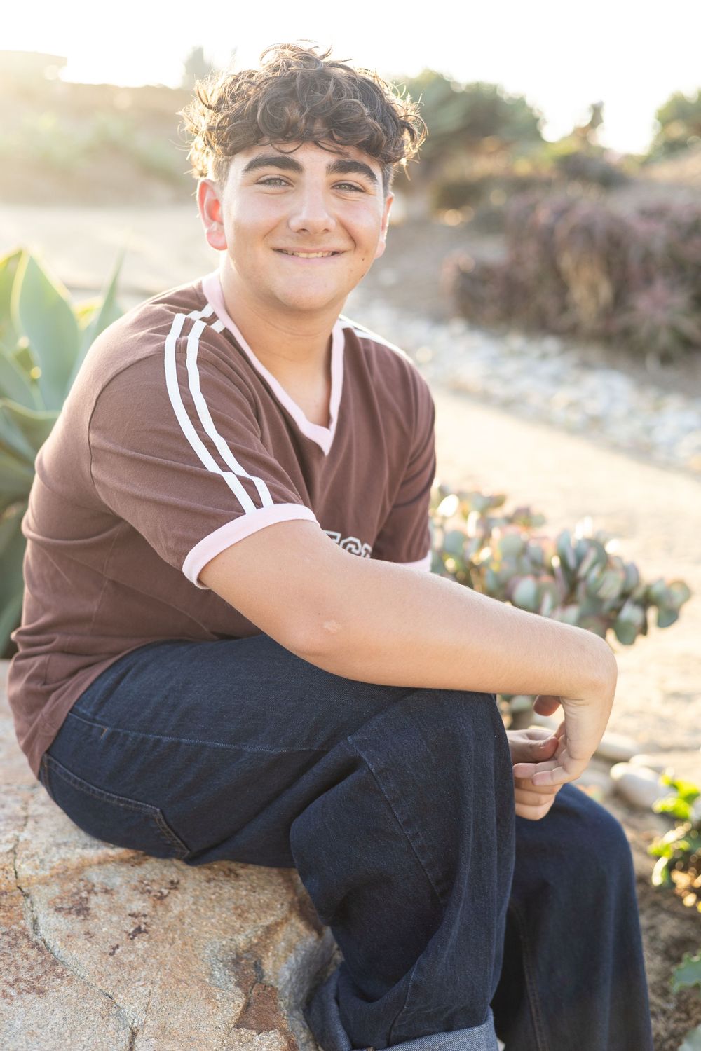 Smiling teenage boy sitting on rocks outdoors in a brown jersey shirt and jeans with golden sunlight behind him.
