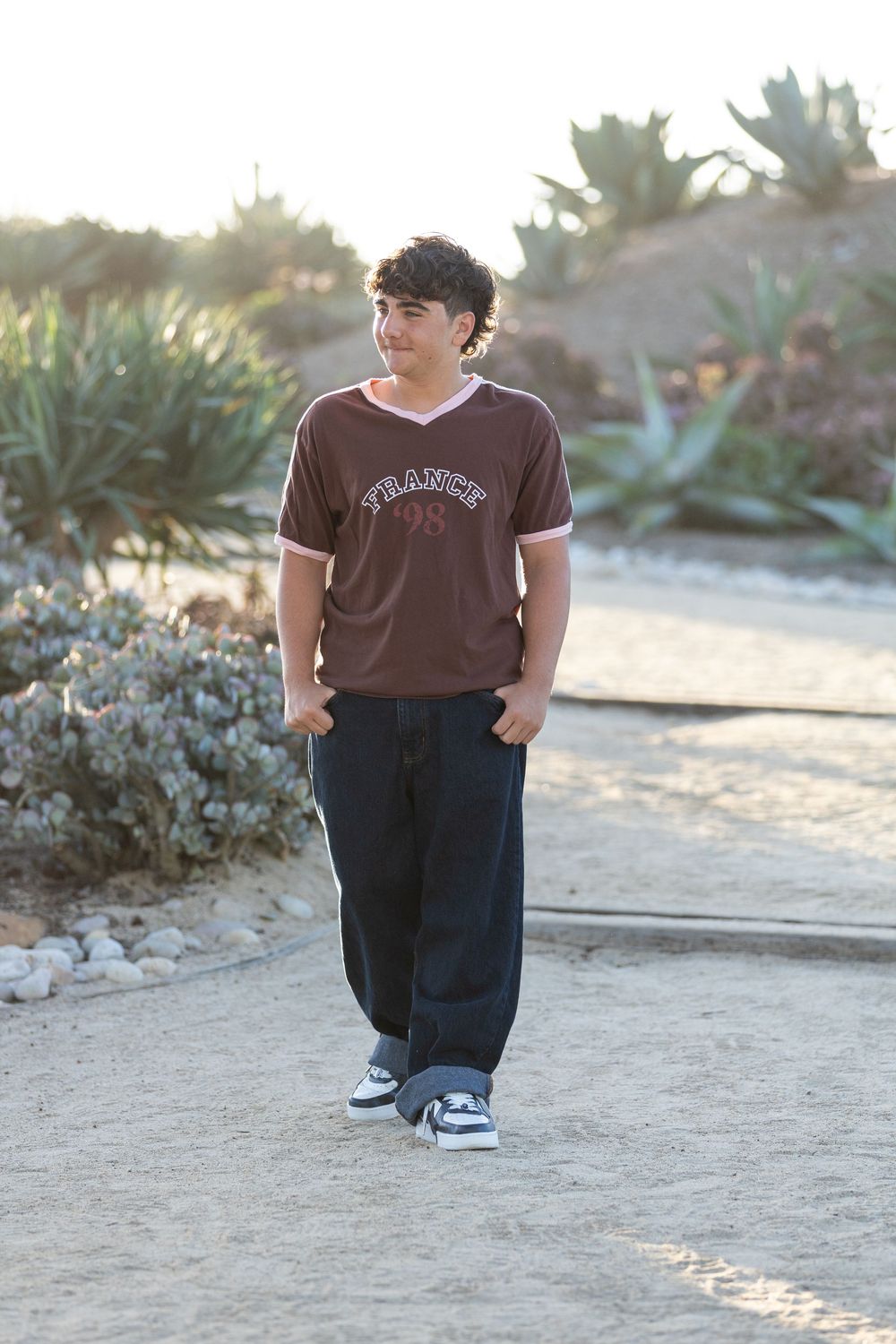 Teen boy in brown France 98 jersey and baggy black pants walking on sandy path with desert plants.