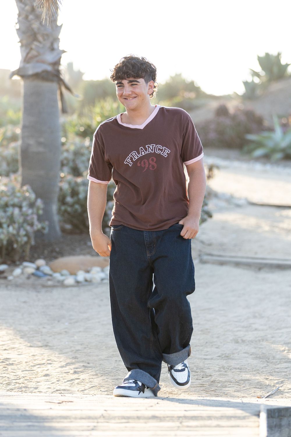 Teen boy walking outdoors in brown France t-shirt, wide-leg jeans, and sneakers on a sunny desert path.