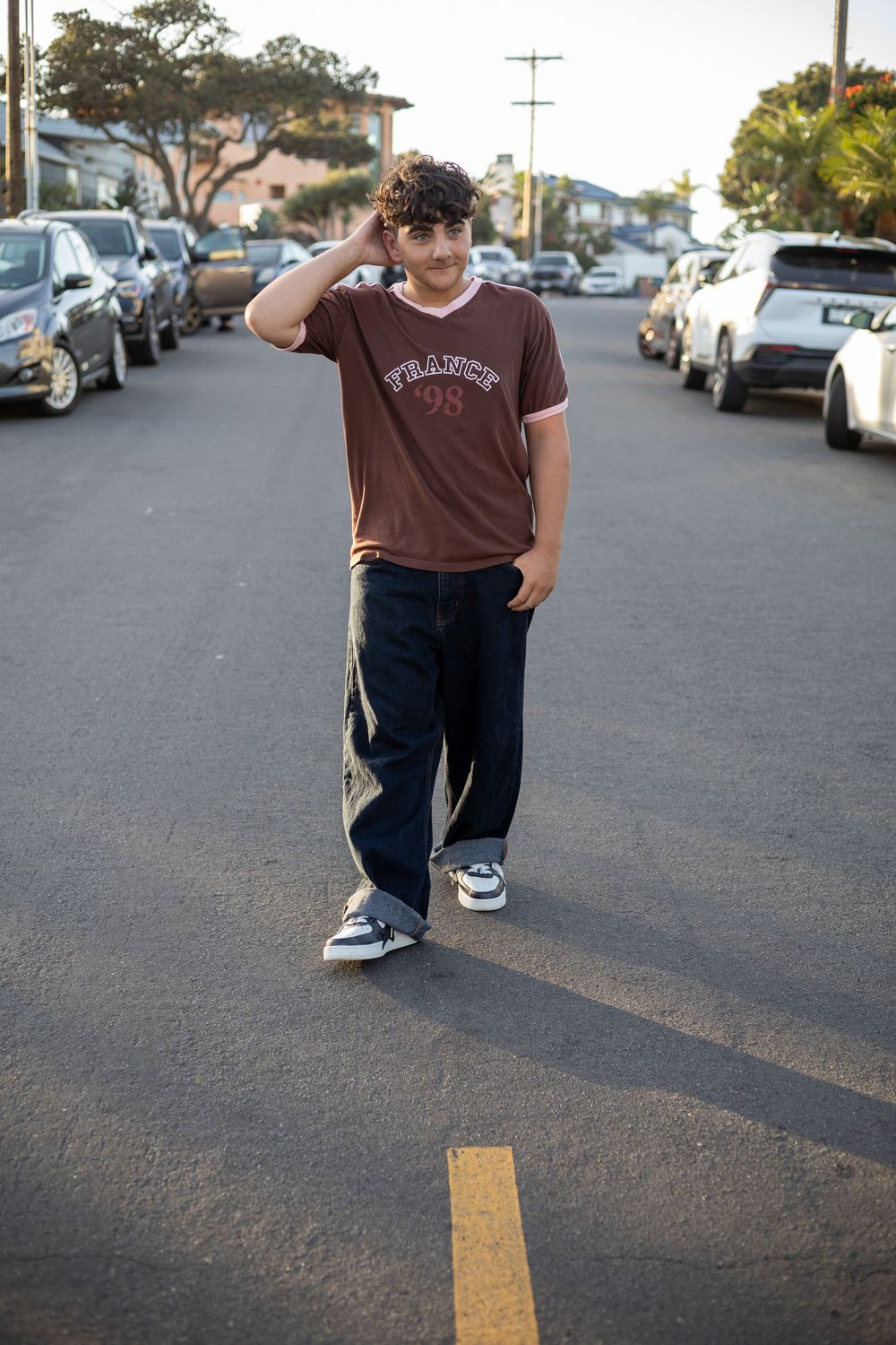 Teen boy in brown France 98 tee and wide-leg jeans poses in parking lot, hand raised to curly hair.