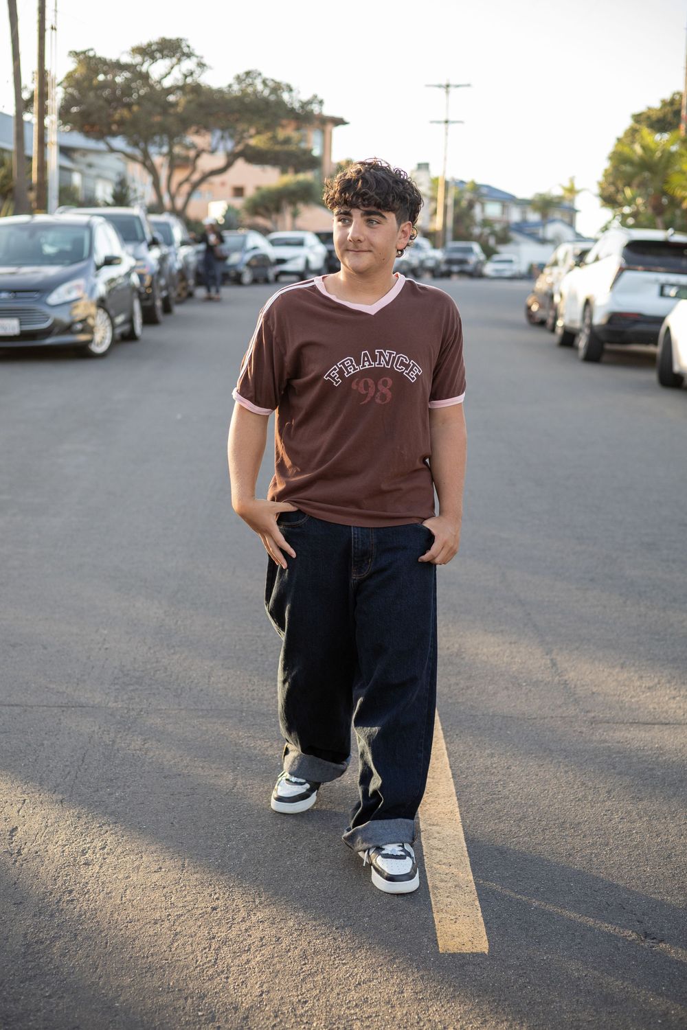 Young man in brown France 98 jersey and navy pants stands confidently in a sunny parking lot.